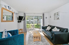A living room with a blue sofa, a black sofa, a wooden coffee table, a TV mounted on the wall, and a large sliding glass door leading to an outdoor area. - Atlantic Reach Cosy 2-bed With Parking (St. Columb Major)