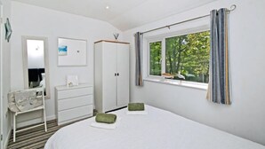 A bedroom with a bed, a dresser, a mirror, a window with curtains, and a view of trees. - Atlantic Reach Cosy 2-bed With Parking (St. Columb Major)