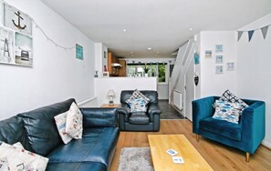 Una sala moderna con un sofá, dos sillones, una mesa de centro y una cocina al fondo.