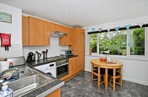 A modern kitchen with wooden cabinets, a stainless steel sink, a microwave, and a stove. There is a dining table with chairs and a window with a view of greenery. - Atlantic Reach Cosy 2-bed With Parking (St. Columb Major)
