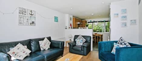 A living room with a sofa, a blue armchair, a wooden coffee table, and a dining area in the background.