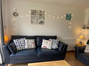 A living room with a black leather sofa, patterned cushions, a wooden coffee table, and string lights. - Atlantic Reach Cosy 2-bed With Parking (St. Columb Major)