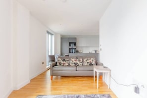 Uma sala de estar moderna com um sofá cinzento, uma mesinha branca e uma cozinha ao fundo.