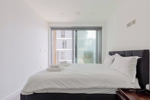 Quarto moderno com cama grande, cabeceira escura e porta de vidro deslizante para uma varanda.