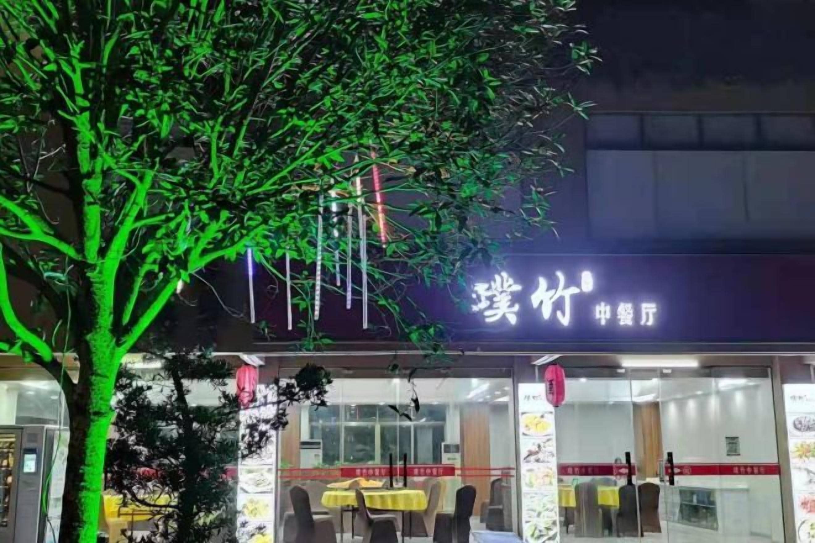 A restaurant with a sign in a foreign language, outdoor seating, and a tree in front.