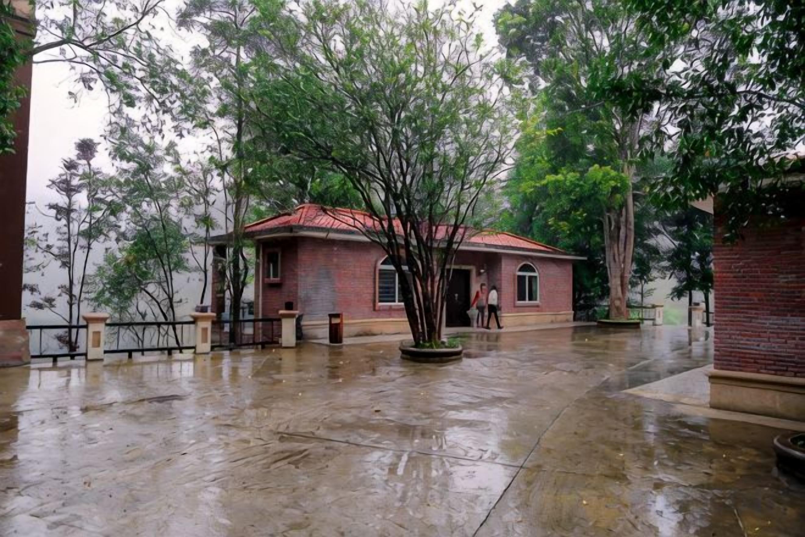 Un edificio de ladrillo con techo rojo, rodeado de árboles y suelo mojado, lo que indica lluvia reciente.