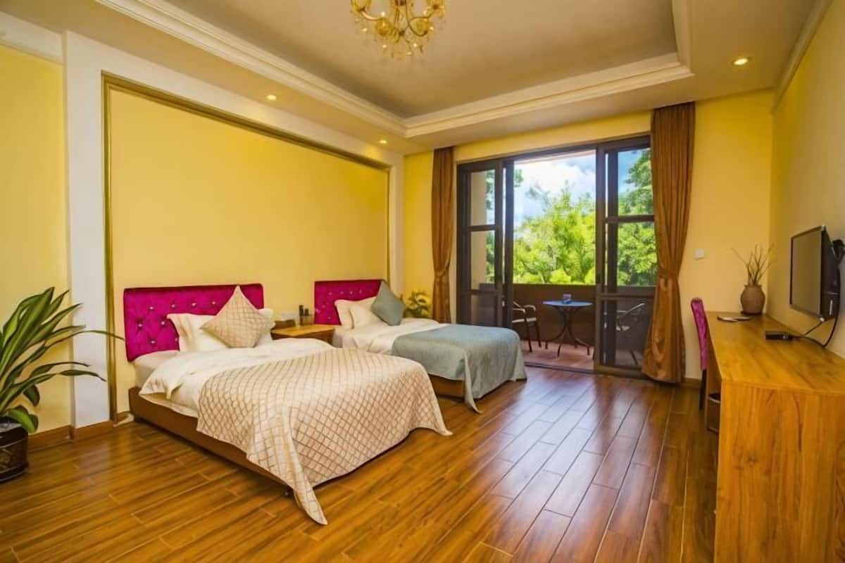 A hotel room with two beds, a wooden floor, a balcony with a table and chairs, and a TV on a wooden cabinet.