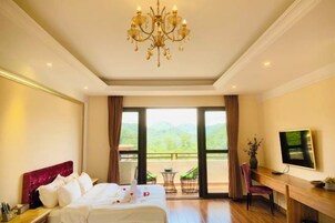 Quarto de hotel com uma cama grande, uma escrivaninha, televisão e varanda com vista para o verde.