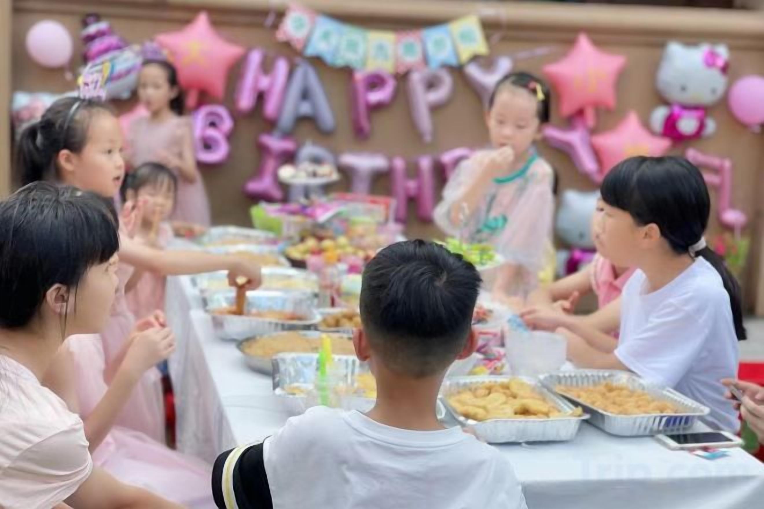 Un grupo de niños celebrando un cumpleaños, con una mesa llena de comida y un cartel que dice '¡Feliz Cumpleaños' al fondo.