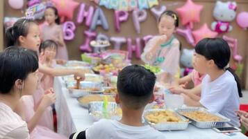 Un grupo de niños celebrando un cumpleaños, con una mesa llena de comida y un cartel que dice '¡Feliz Cumpleaños' al fondo.