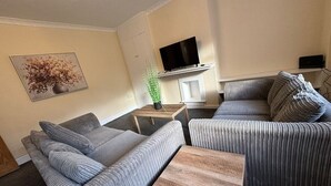 Ein Wohnzimmer mit einer Sofagarnitur, einem Couchtisch, einem Fernseher auf einem Schrank und einer Pflanze auf einem Beistelltisch.