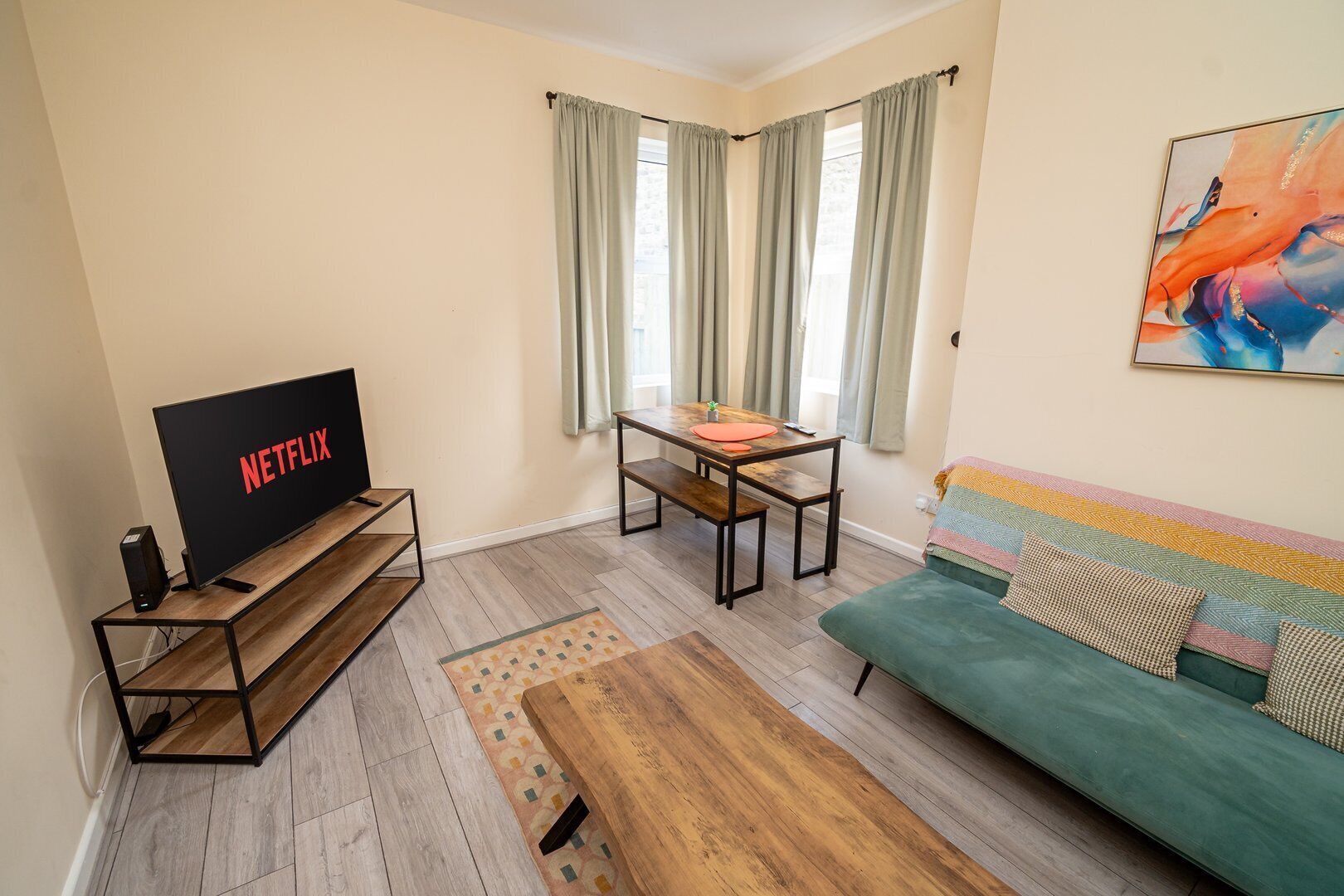 A living room with a TV on a wooden stand, a green sofa, a small table, and a window with curtains.