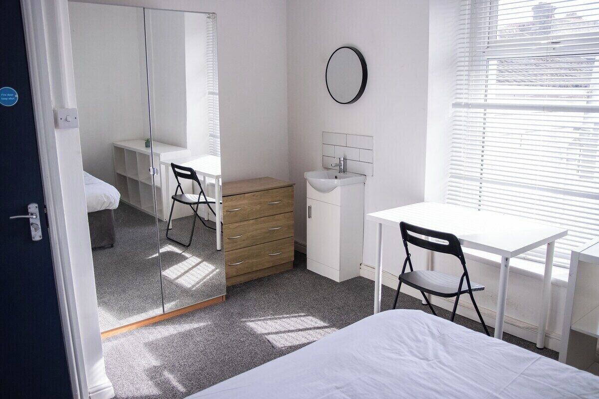 A bedroom with a bed, a desk, a chair, a dresser, a mirror, and a window with blinds.