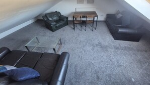 Una camera da letto mansardata con soffitto spiovente, un divano in pelle nera, un tavolino da caffè in vetro, un tavolo da pranzo in legno con sedie e due opere d'arte appese alle pareti.