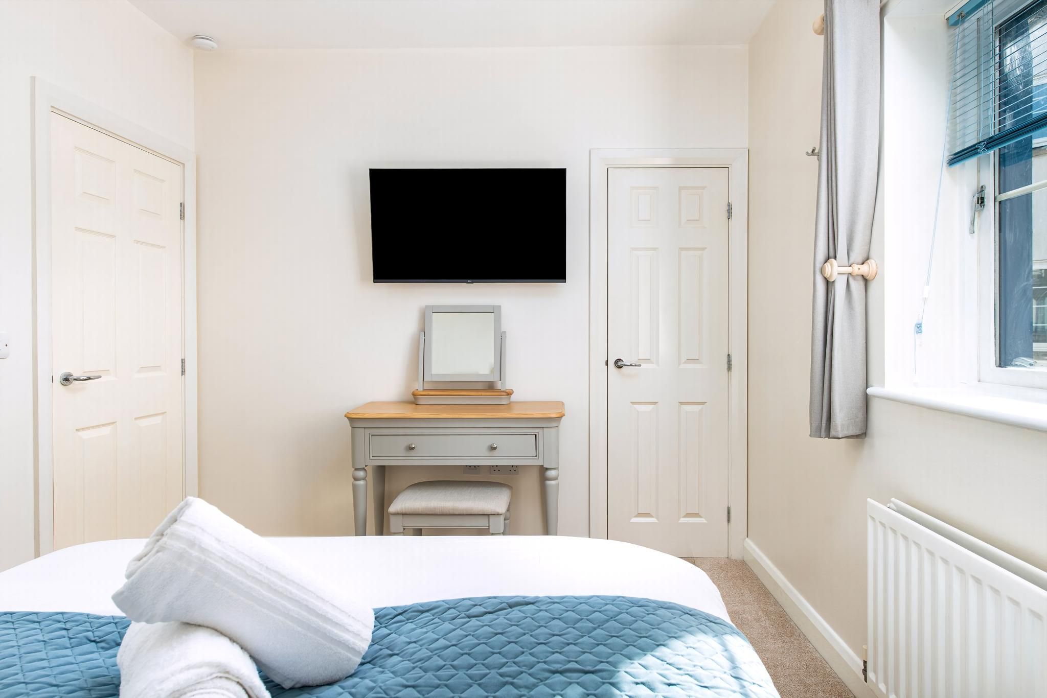 A bedroom with a bed, a television, a desk with a mirror, and a window.