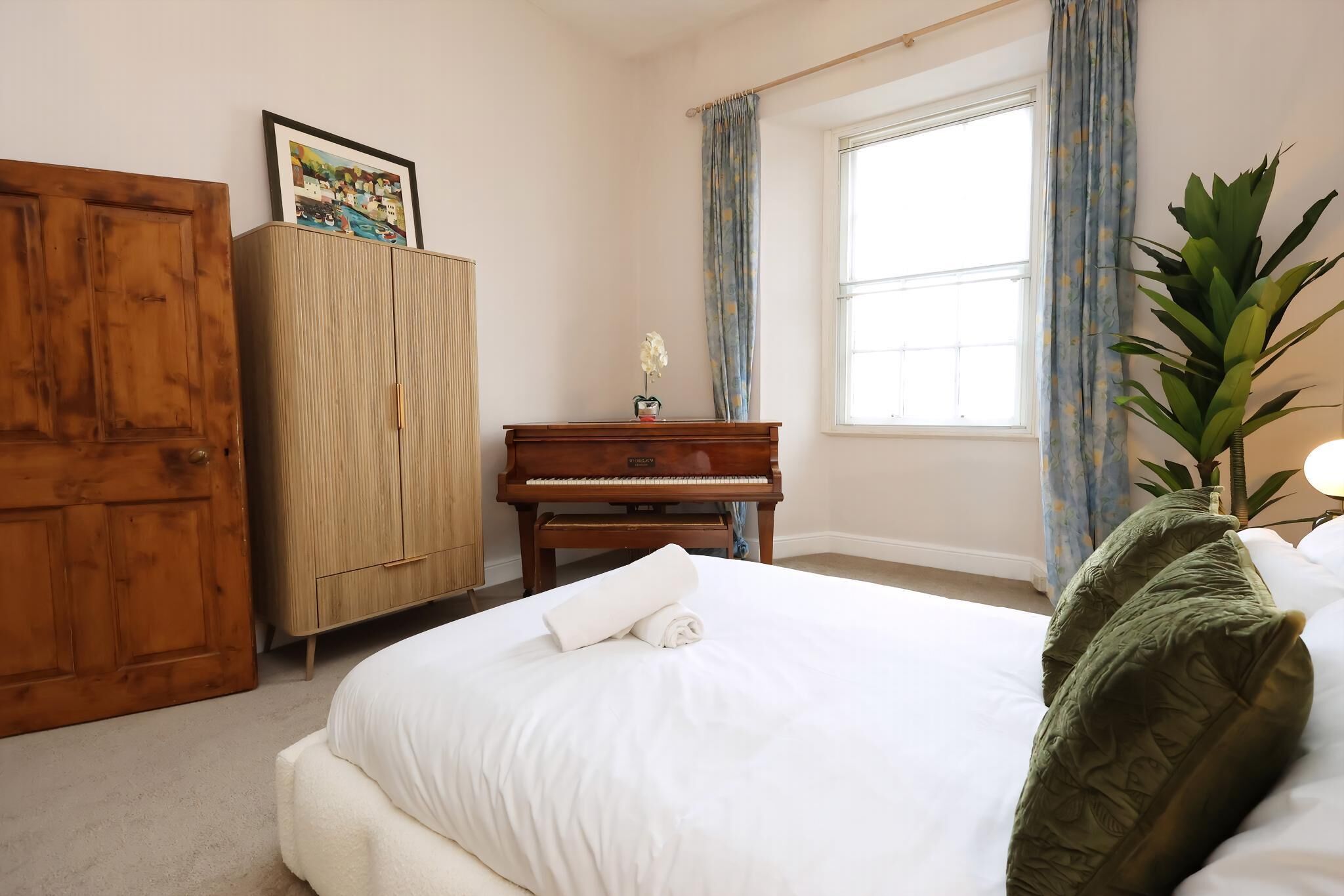 A bedroom with a bed, a wooden wardrobe, a piano, a window with curtains, and a potted plant.