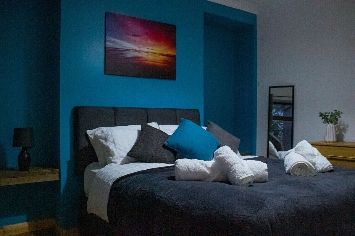 A bedroom with a bed, pillows, a nightstand, a lamp, a painting, and a plant.
