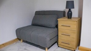 A small, neatly arranged room with a grey sofa bed, a wooden nightstand with a lamp, and a marble-tiled floor. - Spacious & Comfortable 2-bedroom Home (Swansea)