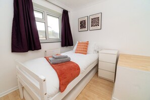 A single-bed room with a white bed, orange and white pillows, a red blanket, a white nightstand, a white dresser, and a window with purple curtains. - Comfortable 3-bed, 2-bath Basingstoke Stay (Basingstoke)