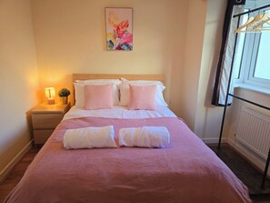 A neatly made bed with pink bedding and two rolled towels, a bedside lamp, and a window with a view. - Bright 4-bed Swansea Getaway With Parking (Swansea)