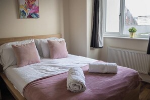 A neatly made bed with pink and white bedding, a rolled white towel, and a window with a view of buildings and a plant on the windowsill. - Bright 4-bed Swansea Getaway With Parking (Swansea)