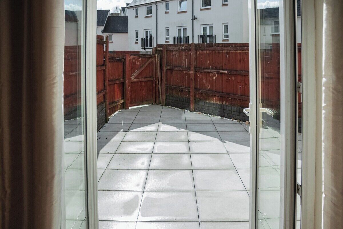A view through a glass door into a courtyard with a tiled floor, a wooden fence, and a building in the background.