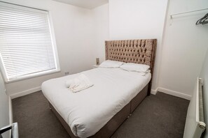 A bedroom with a bed, white bedding, a window with blinds, a radiator, and a wall-mounted heater. - Cosy 3-bed House Walk to Swansea Stadium (Swansea)