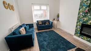 A living room with two dark blue sofas, a dark blue rug, a white radiator, a window with blinds, a small table with a plant, and a wall with a tropical wallpaper. - Cosy 3-bed House Walk to Swansea Stadium (Swansea)