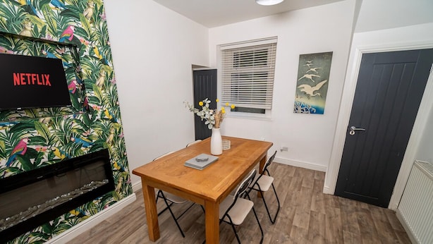 A room with a wooden dining table, chairs, a TV displaying 'NETFLIX,' a fireplace, and a door. - Cosy 3-bed House Walk to Swansea Stadium (Swansea)