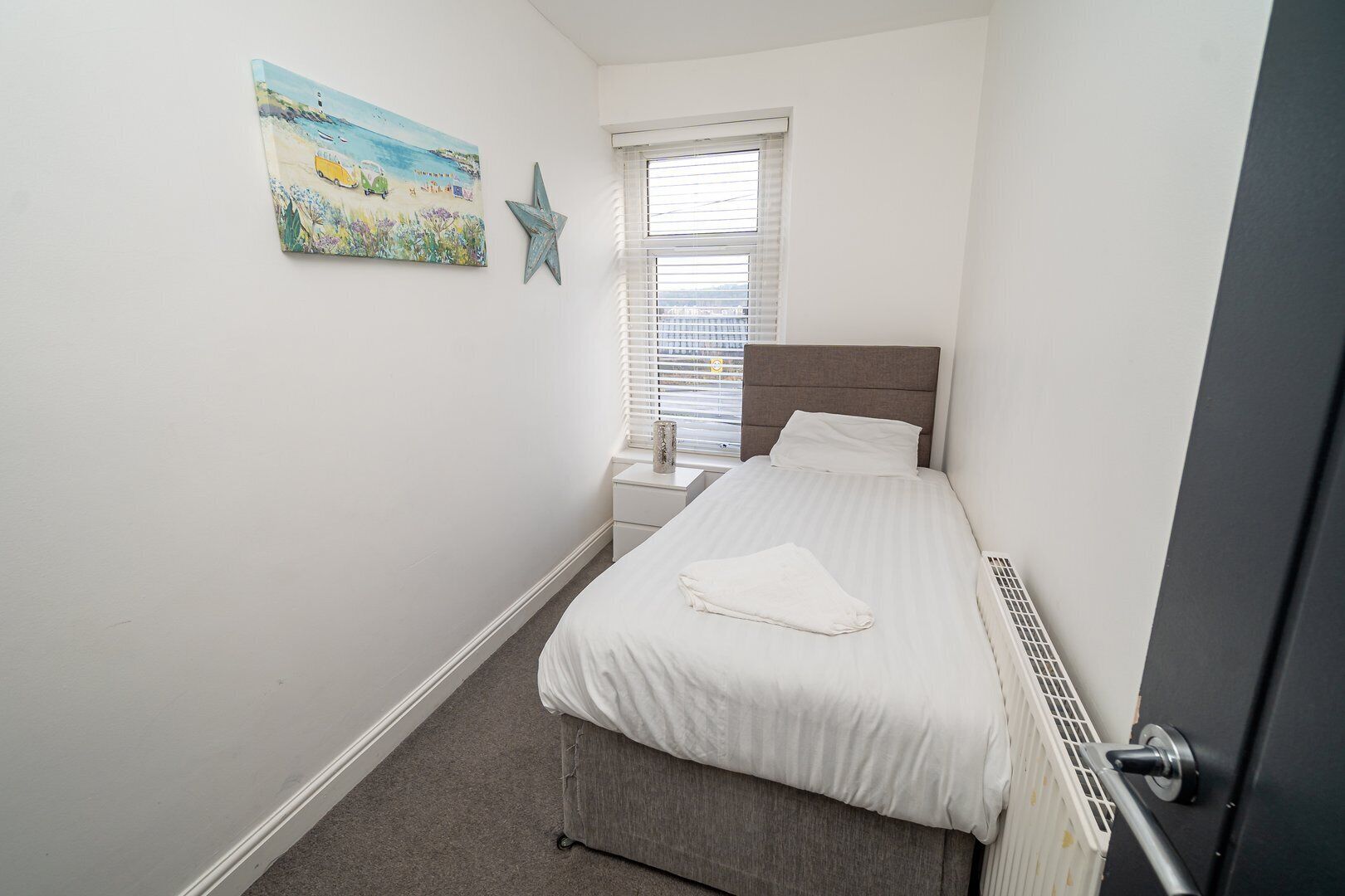 A bedroom with a bed, a nightstand, a window with blinds, a star decoration, and a painting on the wall.