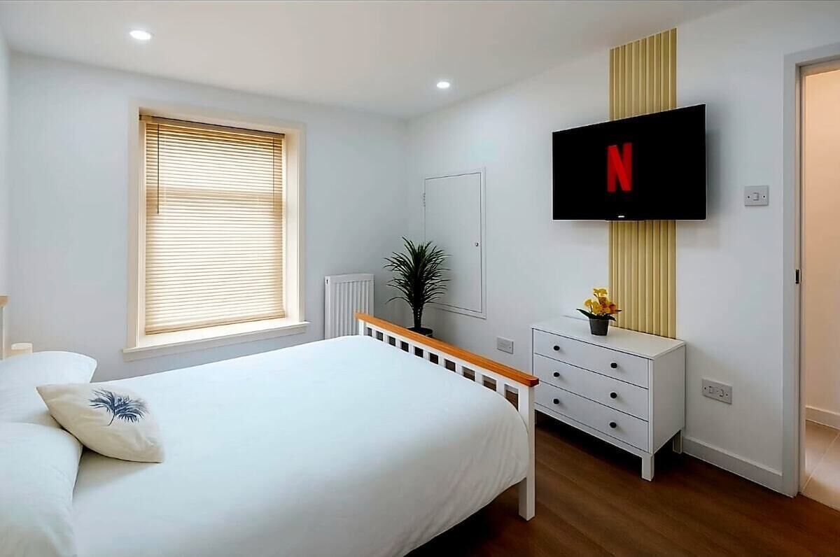 A bedroom with a bed, a TV mounted on the wall, a dresser, and a window with blinds.