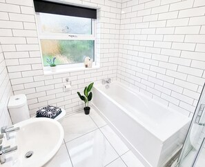 A modern bathroom with white tiled walls, a white bathtub, a sink, and a window with frosted glass. - Spacious 3 Bed Home in the Heart of Swansea (Swansea)
