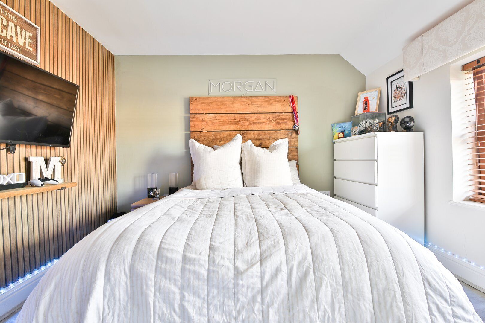 A bedroom with a wooden headboard, a bed with white bedding, a mounted TV, and a white dresser with decorative items.