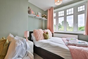 A bedroom with a bed, pillows, a bedside table, a window with curtains, and a shelf with decorative items. - The Swansea Homestay Spacious 4-bed Retreat (Swansea)