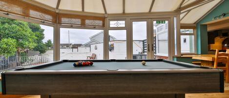 Une table de billard avec ses billes, un banc en bois et une vue sur l’extérieur à travers de grandes fenêtres.
