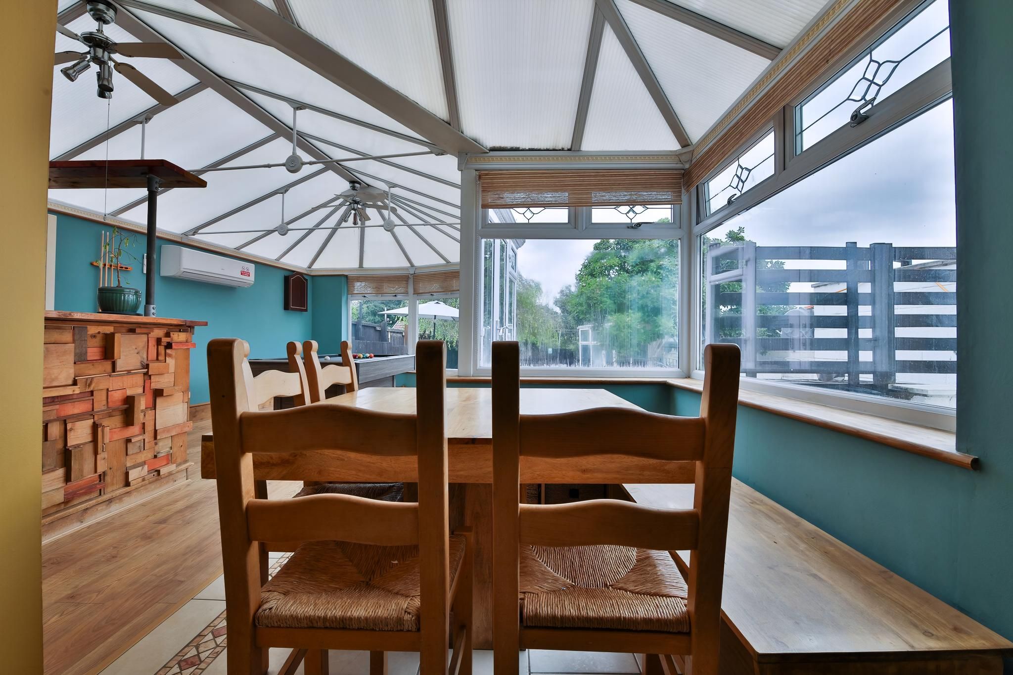 A conservatory with wooden chairs and a table, large windows, and a view of the outside.