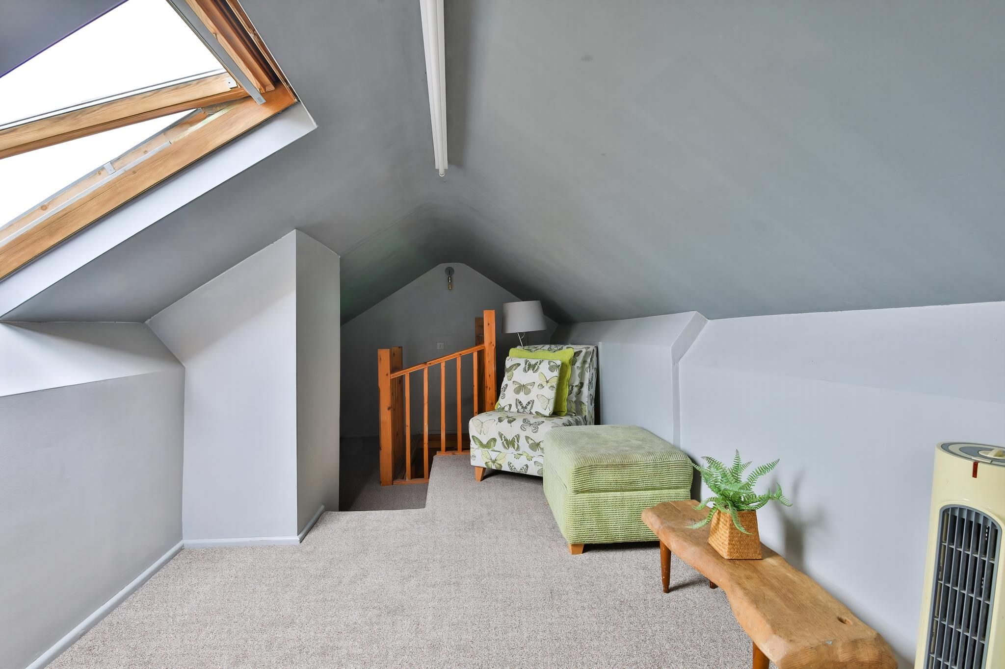 A room with a sloped ceiling, a wooden bench, a green upholstered chair, a small table with a plant, and a radiator.
