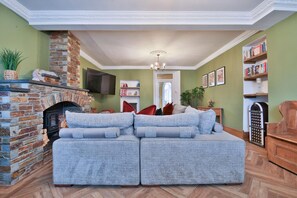 A living room with a stone fireplace, a grey sofa, a television, and a bookshelf. - The Swansea Homestay Spacious 4-bed Retreat (Swansea)