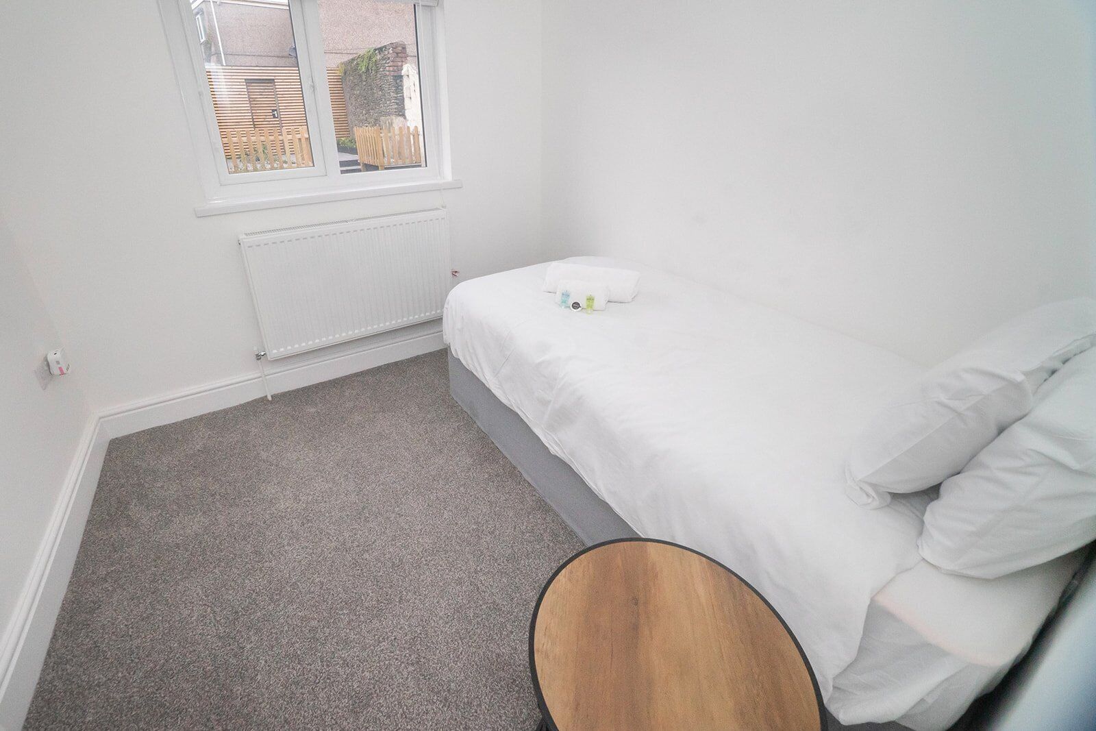 A small, clean room with a single bed, a radiator, a window, and a small round table.
