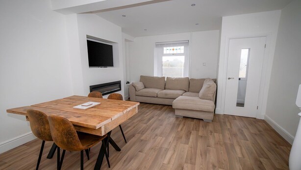 A modern living room with a wooden dining table, two brown chairs, a beige sofa, and a flat-screen TV mounted on the wall. - Spacious 3BR Swansea House Near City Centre (Swansea)