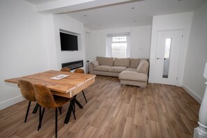 A modern living room with a wooden dining table, two brown chairs, a beige sofa, and a flat-screen TV mounted on the wall. - Spacious 3BR Swansea House Near City Centre (Swansea)
