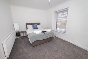 A bedroom with a bed, a bedside table, a mirror, and a window with blinds. - Spacious 3BR Swansea House Near City Centre (Swansea)