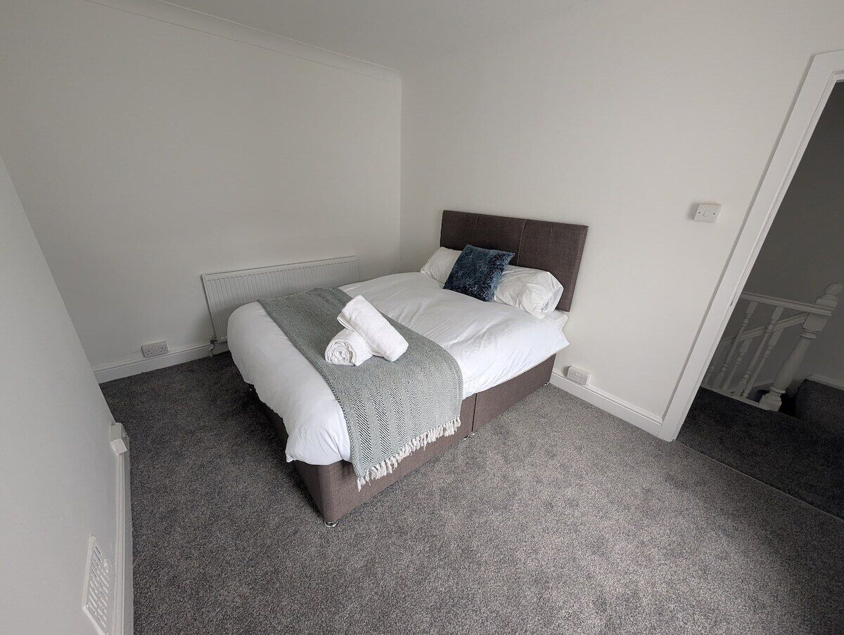 Un dormitorio con una cama, alfombra gris y pared blanca.