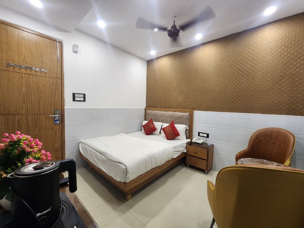A hotel room with a wooden wardrobe, a bed with white bedding and red pillows, a nightstand with a phone, a chair, and a ceiling fan.