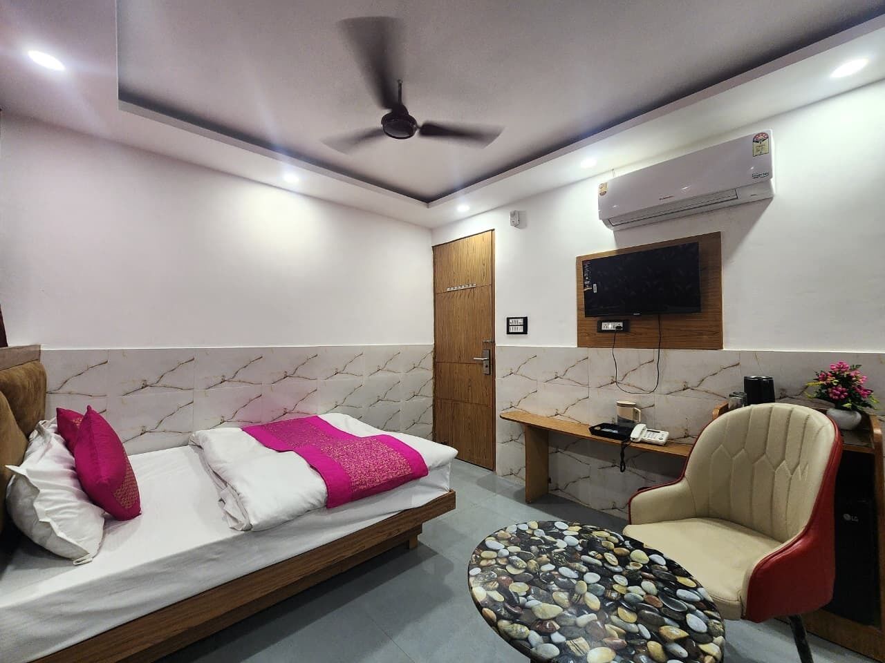 A hotel room with a bed, a desk, a chair, a ceiling fan, an air conditioning unit, and a television.