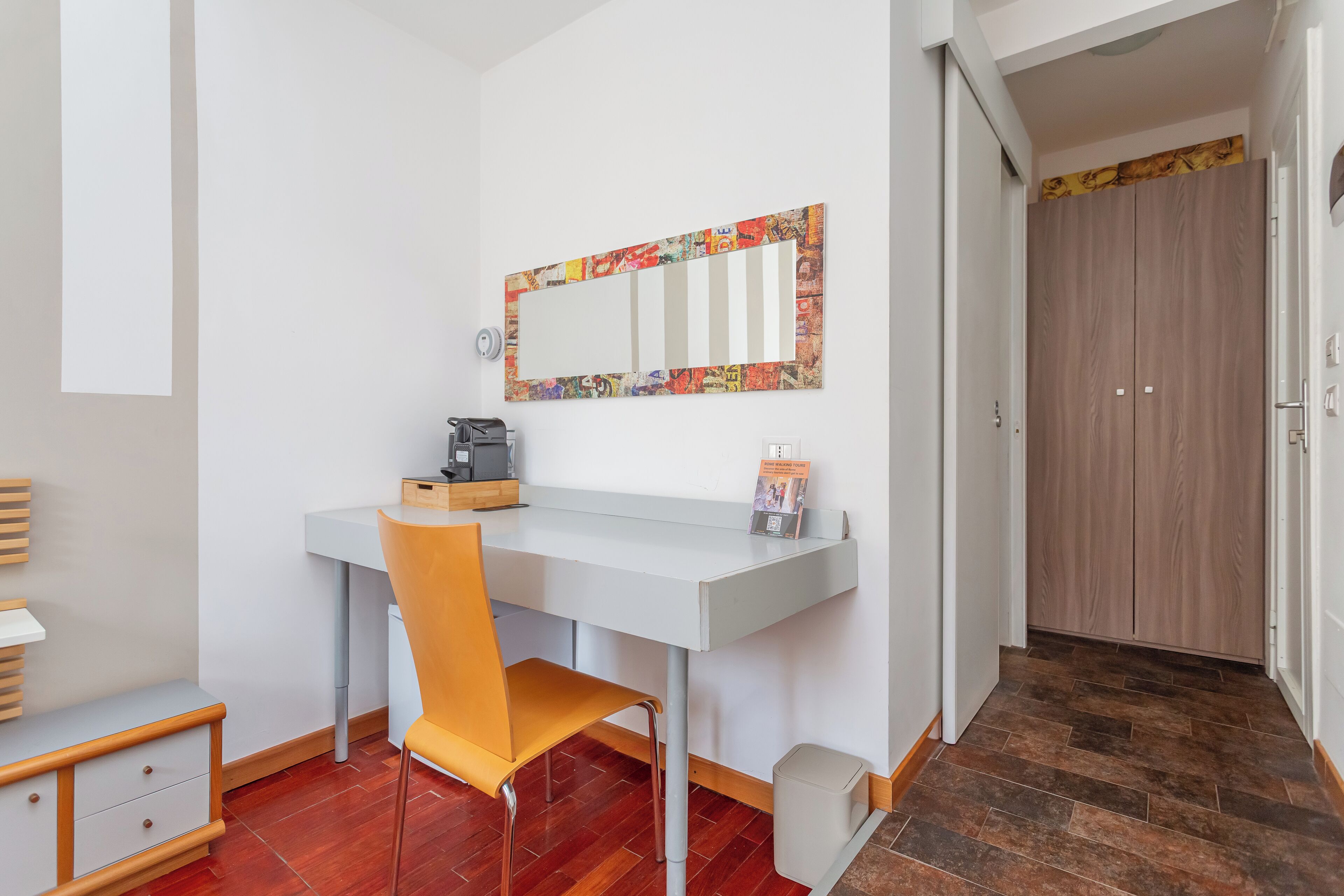 A small, modern room with a white desk, a chair, a mirror with a colorful frame, and a wardrobe.