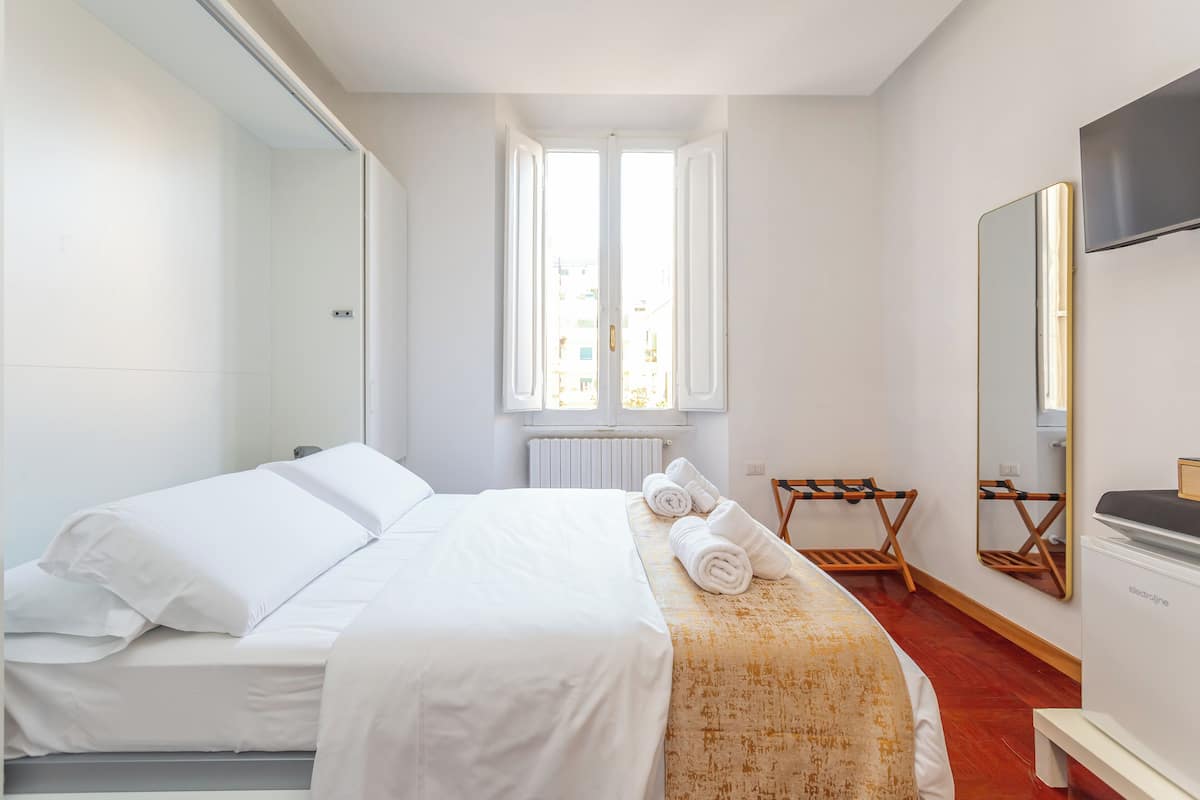 A hotel room with a bed, a wooden bench, a mirror, and a window with white curtains.