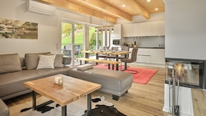 A modern living room with a sofa, a wooden coffee table, and a dining area with a table and chairs. There is a kitchen area with cabinets and a fireplace. - Penthouse Cassiopeia (Tauplitz)