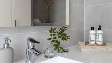 A modern bathroom with a white sink, chrome faucet, and a large mirror. There are two bottles of oil on the counter, and a small plant beside the sink.