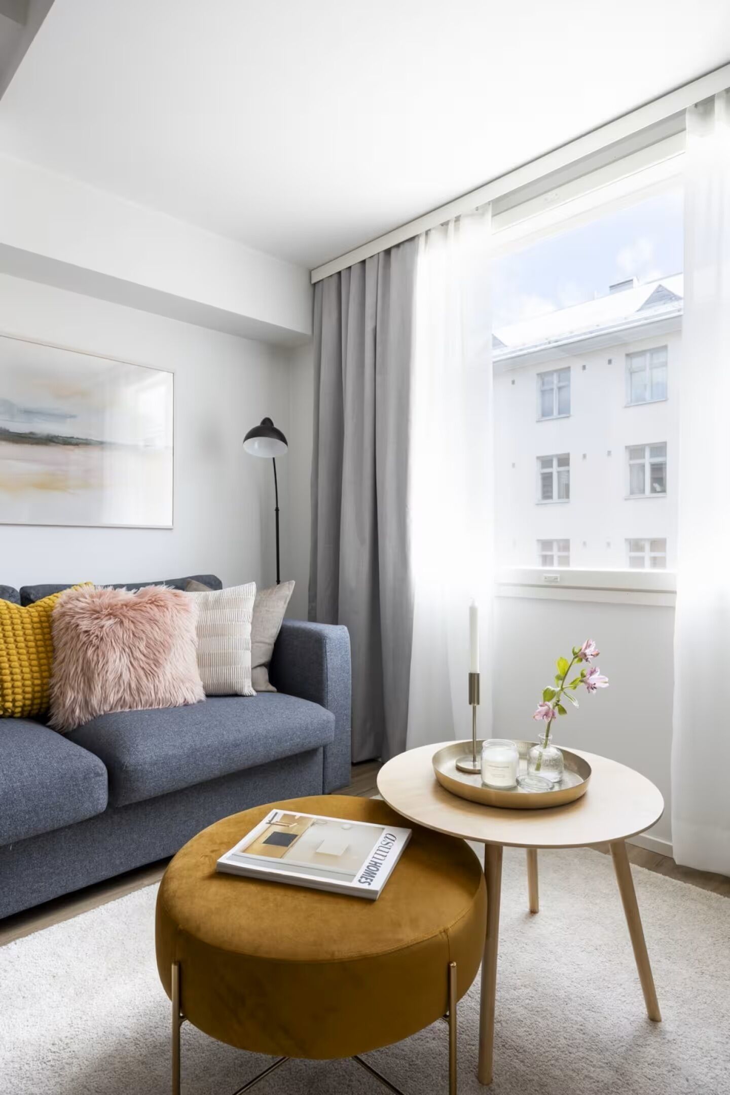 Ein modernes Wohnzimmer mit einem grauen Sofa, einem runden Hocker, einem kleinen Tisch mit Kerze und Blumen und einem großen Fenster mit Vorhängen.